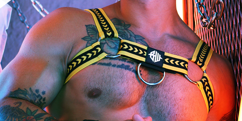 Person wearing a yellow and black HARNESS with tattoos on chest and arms.