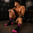 A shirtless man sitting on a gym floor in front of black lockers, wearing black athletic socks with bright pink toes, heels, and cuffs. The socks have the brand name 'PUP' printed in white on the cuffs.